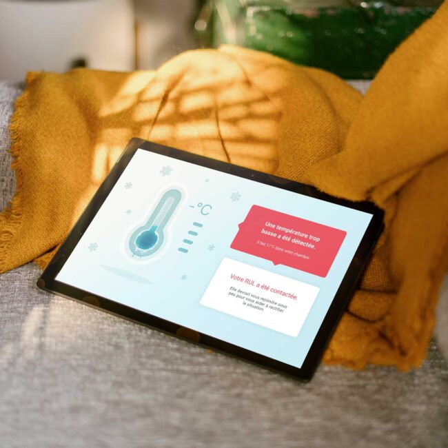 Image présentant une tablette posée sur un une couverture sur un sofa. La tablette affiche un projet d'interactions ambiantes du laboratoire Domus : un thermomètre glacé avec un avertissement disant : "Une température trop basse a été détectée. Il fait 17 degrés celsius dans votre chambre. Votre Responsable d'unité de logement a été contactée. Elle devrait vous rejoindre sous peu vous vous aider à rectifier la situation".